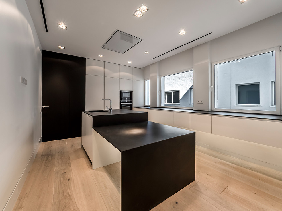 Design ideas for a mid-sized modern u-shaped separate kitchen in Madrid with an undermount sink, flat-panel cabinets, white cabinets, panelled appliances, light hardwood floors, with island and black benchtop.