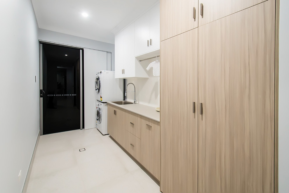 This is an example of a mid-sized modern single-wall dedicated laundry room in Other with a drop-in sink, flat-panel cabinets, light wood cabinets, laminate benchtops, white splashback, ceramic splashback, white walls, ceramic floors, a stacked washer and dryer, white floor and beige benchtop.