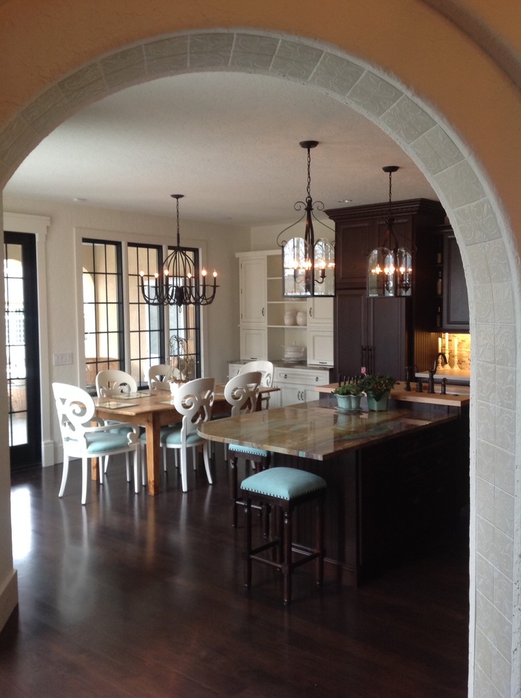 This is an example of a mid-sized mediterranean l-shaped eat-in kitchen in Other with a farmhouse sink, raised-panel cabinets, dark wood cabinets, granite benchtops, panelled appliances, dark hardwood floors and with island.