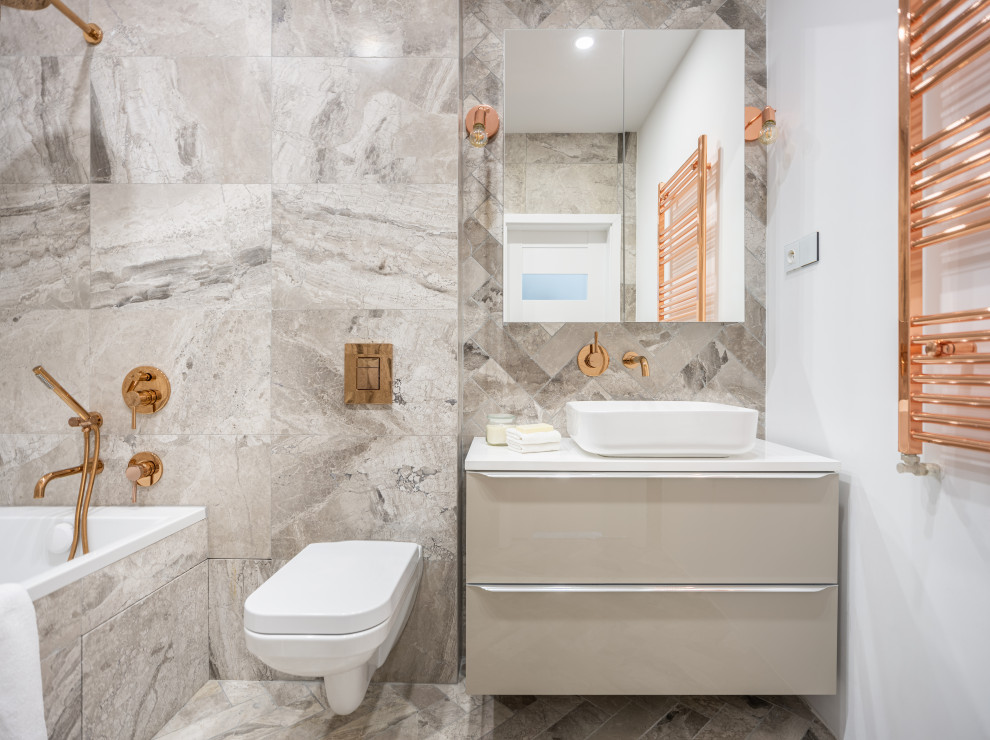 Small contemporary grey and cream ensuite bathroom in London with flat-panel cabinets, beige cabinets, a built-in bath, a wall mounted toilet, brown tiles, stone tiles, multi-coloured walls, ceramic flooring, a vessel sink, brown floors, a feature wall, a single sink, a floating vanity unit and a drop ceiling.