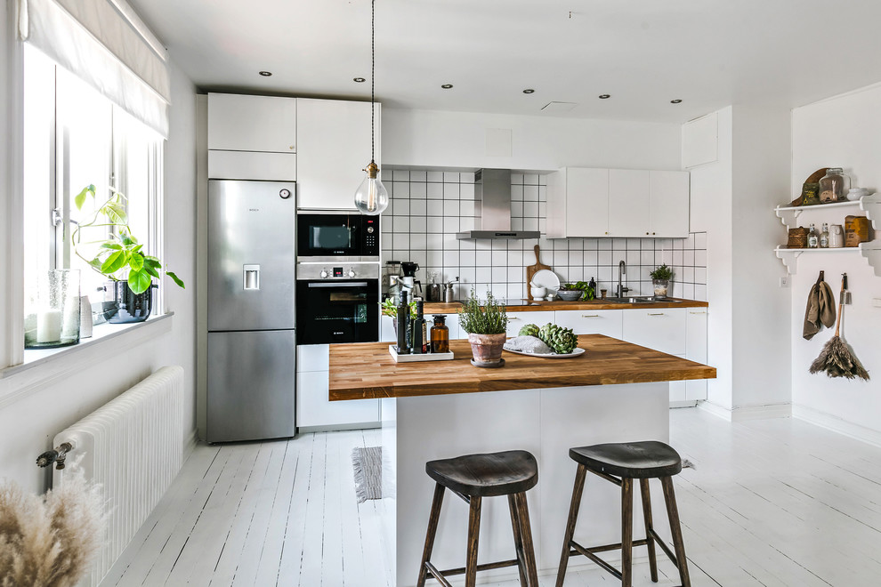 This is an example of a scandinavian single-wall kitchen in Gothenburg with a drop-in sink, flat-panel cabinets, white cabinets, wood benchtops, white splashback, stainless steel appliances, painted wood floors, with island, white floor and brown benchtop.