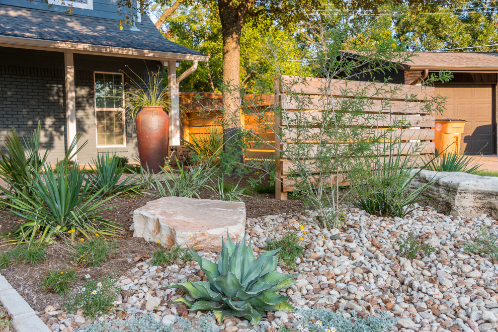 This is an example of a mid-sized and desert look contemporary front yard partial sun xeriscape for summer in Austin with a wood fence and river rock.