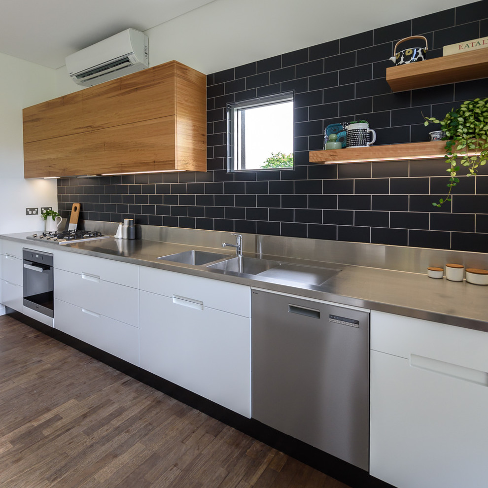 Inspiration for a mid-sized modern galley open plan kitchen in Melbourne with an integrated sink, flat-panel cabinets, white cabinets, stainless steel benchtops, black splashback, ceramic splashback, stainless steel appliances, medium hardwood floors, with island and brown floor.