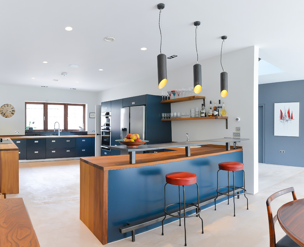 Design ideas for a mid-sized contemporary l-shaped open plan kitchen in Sussex with a drop-in sink, flat-panel cabinets, blue cabinets, wood benchtops, black splashback, ceramic splashback, concrete floors, with island and stainless steel appliances.