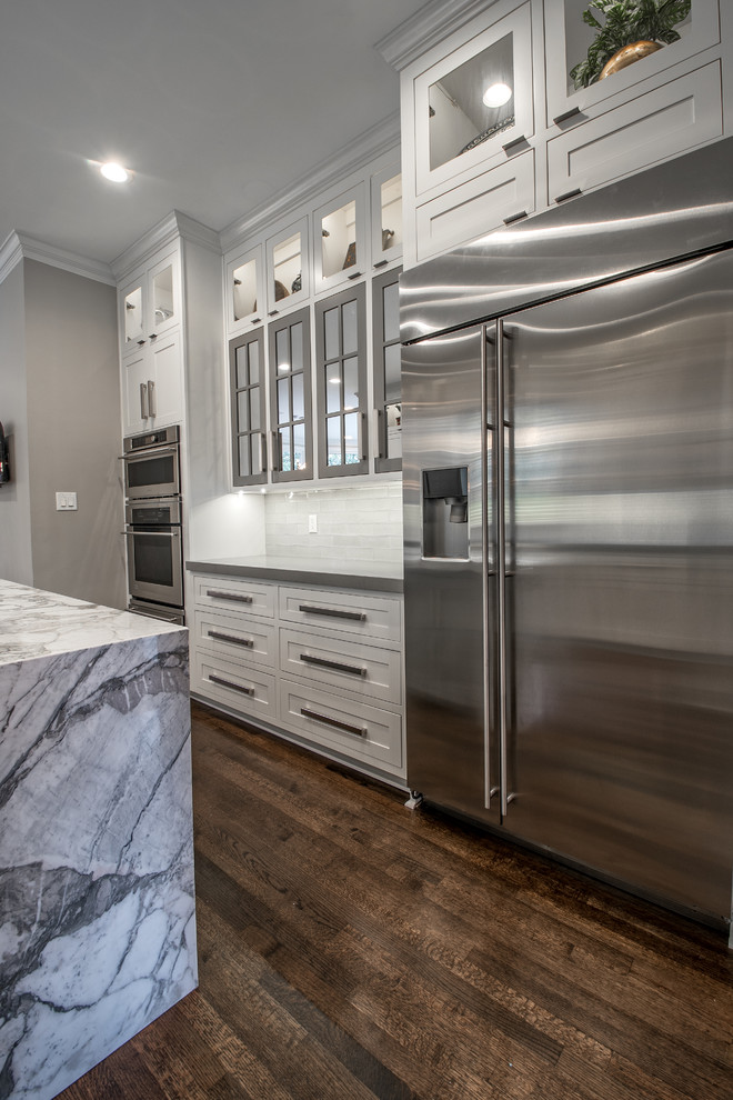 Enclosed kitchen - large transitional l-shaped dark wood floor enclosed kitchen idea with a farmhouse sink, shaker cabinets, white cabinets, solid surface countertops, white backsplash, subway tile backsplash, stainless steel appliances and an island