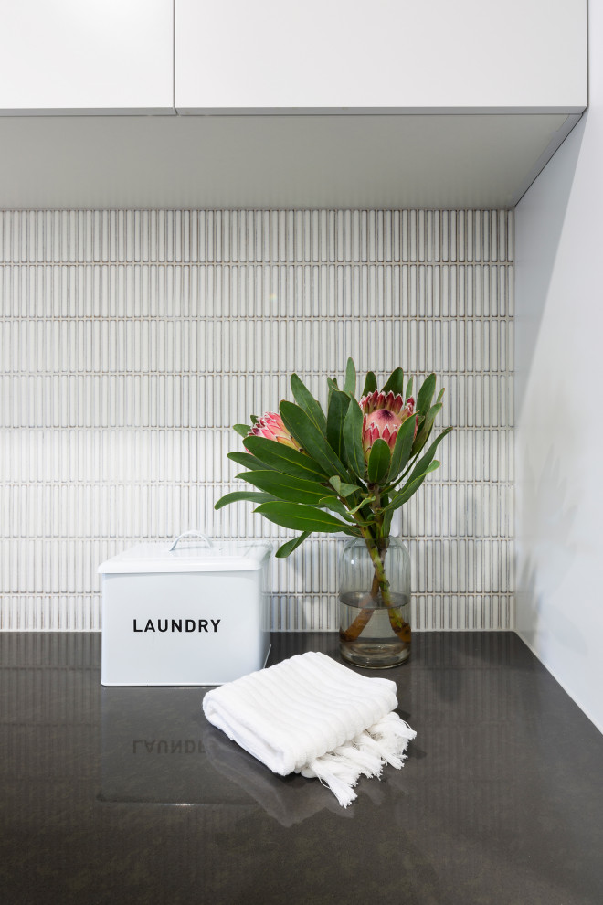 Design ideas for a contemporary single-wall laundry room in Canberra - Queanbeyan with a drop-in sink, white cabinets, quartz benchtops, white splashback, mosaic tile splashback, white walls, porcelain floors, a side-by-side washer and dryer, grey floor and black benchtop.