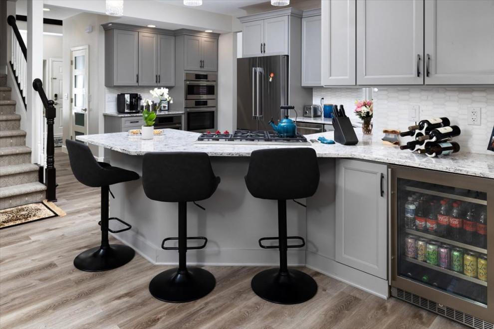 Example of a mid-sized transitional laminate floor and gray floor open concept kitchen design with recessed-panel cabinets, gray cabinets, quartz countertops, white backsplash, marble backsplash, stainless steel appliances and white countertops