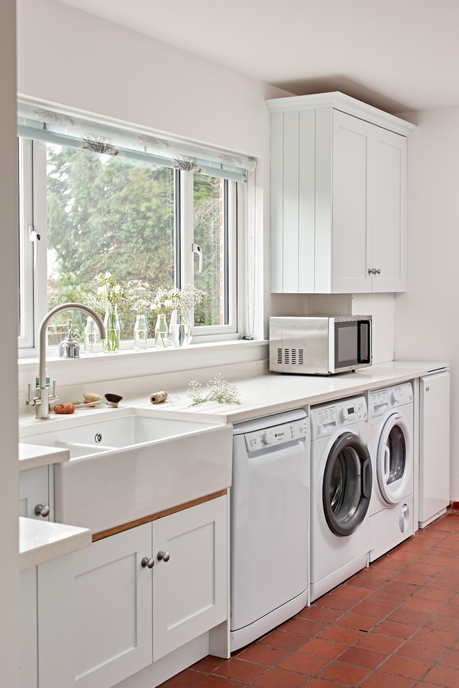 This is an example of a mid-sized transitional dedicated laundry room in Wiltshire with a farmhouse sink, recessed-panel cabinets, white cabinets, terra-cotta floors, a side-by-side washer and dryer and red floor.