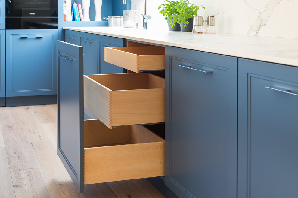 Mittelgroße Klassische Küche mit integriertem Waschbecken, Schrankfronten im Shaker-Stil, blauen Schränken, Küchenrückwand in Weiß, schwarzen Elektrogeräten, braunem Holzboden, Kücheninsel, braunem Boden und weißer Arbeitsplatte in München