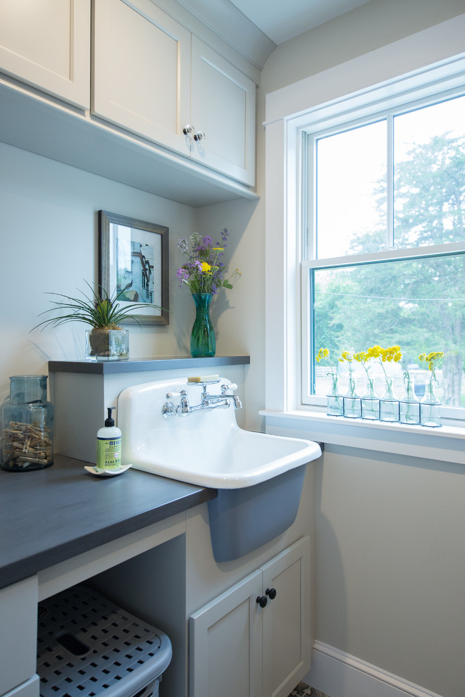 Photo of a beach style galley dedicated laundry room in Boston with a farmhouse sink, shaker cabinets, white cabinets, solid surface benchtops, beige walls, ceramic floors, a side-by-side washer and dryer, multi-coloured floor and grey benchtop.