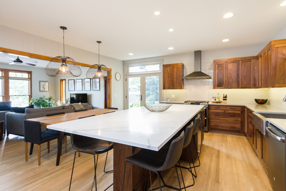 Mid-sized contemporary l-shaped eat-in kitchen in Kansas City with a farmhouse sink, recessed-panel cabinets, medium wood cabinets, quartz benchtops, white splashback, ceramic splashback, stainless steel appliances, light hardwood floors, with island and white benchtop.