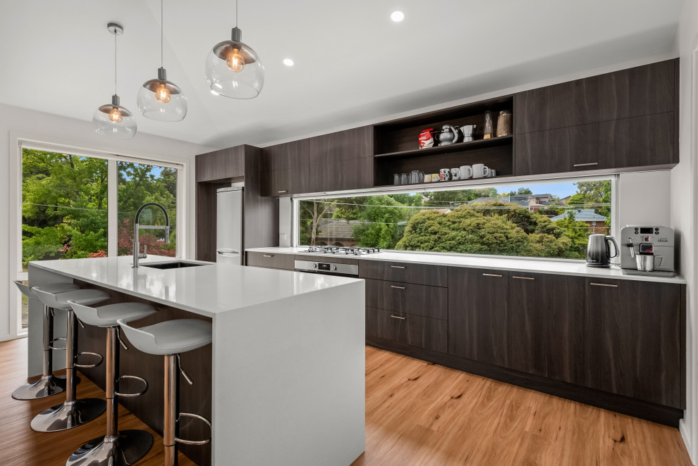Mid-sized modern galley open plan kitchen in Melbourne with a drop-in sink, flat-panel cabinets, dark wood cabinets, granite benchtops, window splashback, stainless steel appliances, medium hardwood floors, with island, white benchtop and wallpaper.