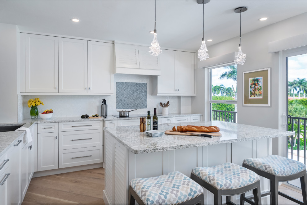 Beach style grey and white l-shaped kitchen in Miami with a built-in sink, flat-panel cabinets, white cabinets, white splashback, light hardwood flooring, an island, brown floors and grey worktops.