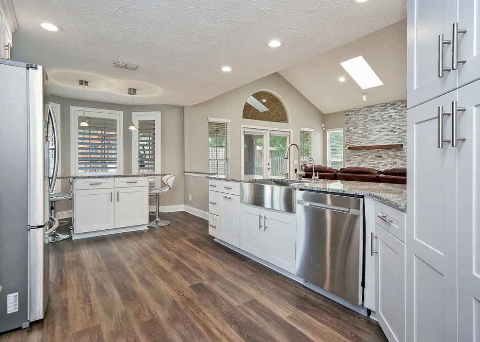 Photo of a mid-sized transitional u-shaped kitchen in Jacksonville with a farmhouse sink, recessed-panel cabinets, white cabinets, granite benchtops, grey splashback, slate splashback, stainless steel appliances, medium hardwood floors, brown floor and grey benchtop.