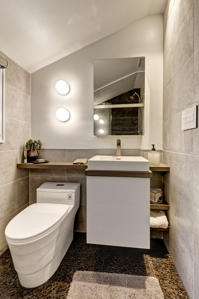 Inredning av ett modernt litet brun brunt badrum med dusch, med släta luckor, vita skåp, en dusch i en alkov, en toalettstol med hel cisternkåpa, grå kakel, porslinskakel, grå väggar, mosaikgolv, ett avlångt handfat, träbänkskiva, brunt golv och dusch med skjutdörr