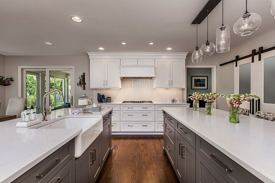 Design ideas for a large traditional grey and white galley kitchen/diner in Detroit with a belfast sink, shaker cabinets, grey cabinets, engineered stone countertops, white splashback, porcelain splashback, stainless steel appliances, medium hardwood flooring, multiple islands, brown floors and white worktops.