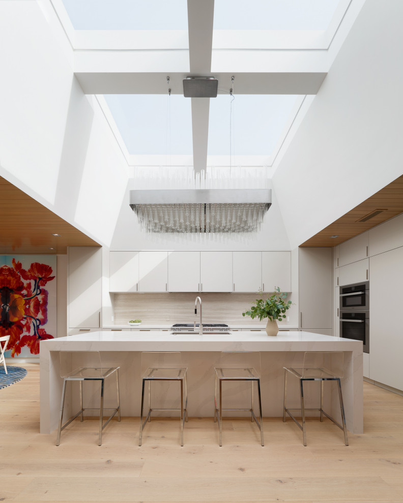 Inspiration for a contemporary l-shaped kitchen in Los Angeles with a submerged sink, flat-panel cabinets, white cabinets, beige splashback, stainless steel appliances, light hardwood flooring, an island, beige floors and white worktops.