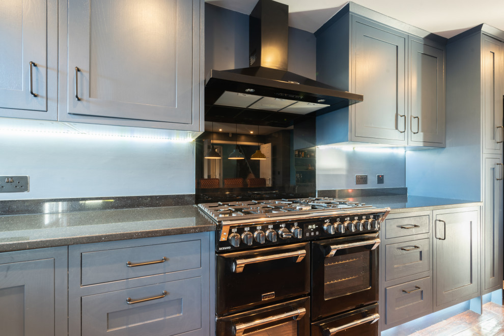 Inspiration for a large contemporary eat-in kitchen in Kent with a farmhouse sink, shaker cabinets, blue cabinets, quartzite benchtops, black splashback, glass sheet splashback, black appliances, dark hardwood floors, with island, brown floor and black benchtop.