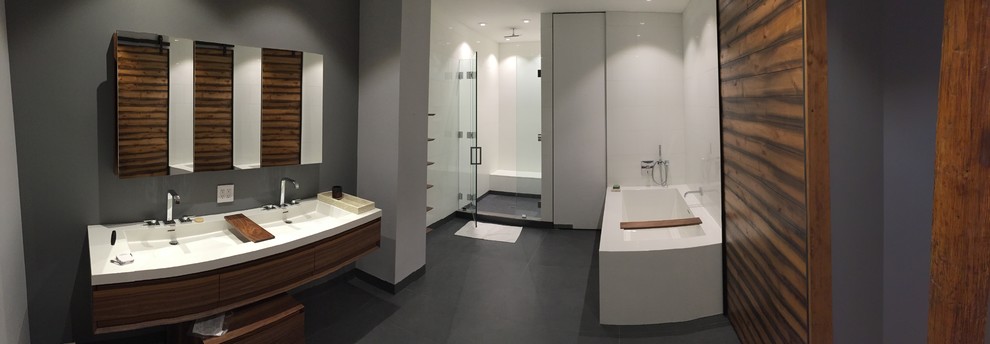 This is an example of a large industrial master bathroom in New York with flat-panel cabinets, medium wood cabinets, a drop-in tub, an alcove shower, a two-piece toilet, grey walls, cement tiles, a drop-in sink, solid surface benchtops, grey floor, a hinged shower door and white benchtops.