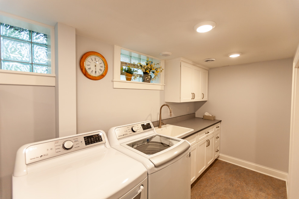 Transitional single-wall dedicated laundry room in Minneapolis with a single-bowl sink, recessed-panel cabinets, white cabinets, solid surface benchtops, grey walls, linoleum floors, a side-by-side washer and dryer, beige floor and grey benchtop.