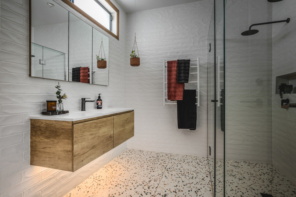 This is an example of a medium sized midcentury ensuite bathroom in Melbourne with medium wood cabinets, a double shower, a wall mounted toilet, white tiles, ceramic tiles, white walls, terrazzo flooring, a submerged sink, engineered stone worktops, multi-coloured floors, a hinged door, white worktops, a single sink and a floating vanity unit.