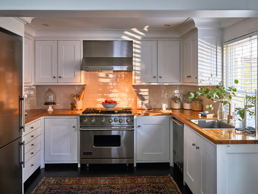 This is an example of a mid-sized transitional u-shaped separate kitchen in Providence with a drop-in sink, shaker cabinets, white cabinets, wood benchtops, white splashback, subway tile splashback, stainless steel appliances, painted wood floors and black floor.