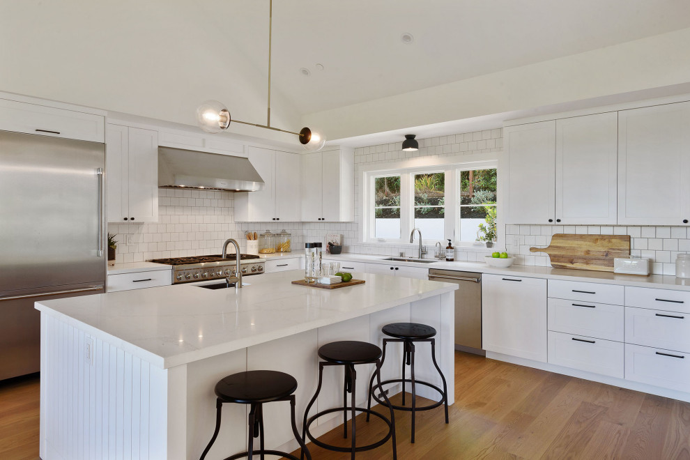 Large transitional l-shaped kitchen in San Francisco with an undermount sink, recessed-panel cabinets, white cabinets, white splashback, stainless steel appliances, with island, brown floor and white benchtop.
