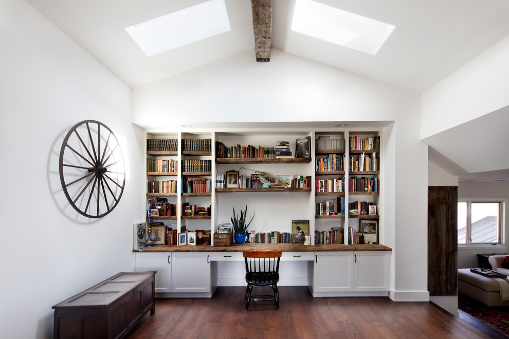 Country home office in Austin with a library, white walls, medium hardwood floors, a built-in desk and brown floor.