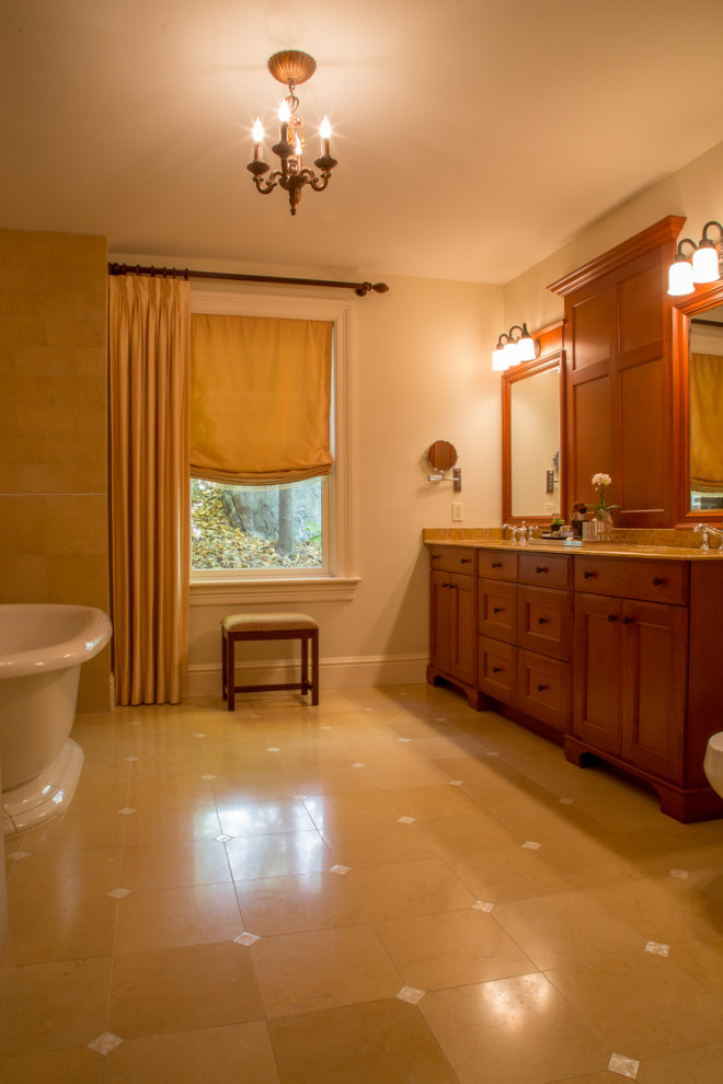 Photo of a large ensuite bathroom in New York with freestanding cabinets, medium wood cabinets, a freestanding bath, an alcove shower, a bidet, beige tiles, limestone tiles, beige walls, limestone flooring, a built-in sink, marble worktops, beige floors, a hinged door, beige worktops, double sinks and a built in vanity unit.