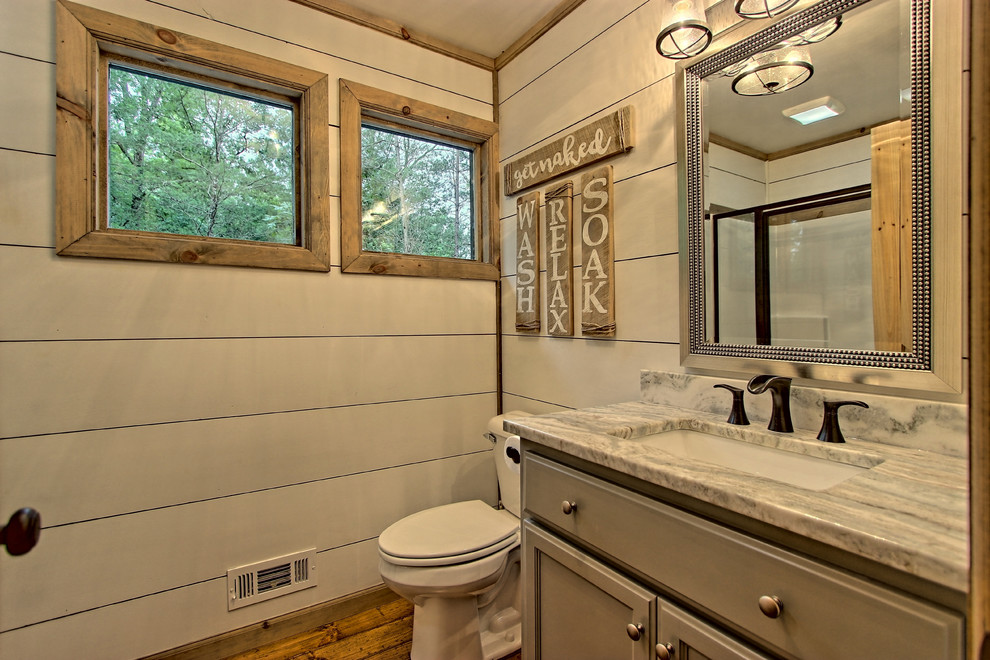 Photo of a small country bathroom in Other with recessed-panel cabinets, grey cabinets, a two-piece toilet, white walls, painted wood floors, a drop-in sink, granite benchtops, brown floor and a hinged shower door.