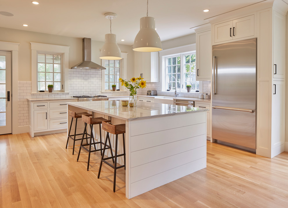 Inspiration for a transitional l-shaped medium tone wood floor and brown floor kitchen remodel in Boston with a farmhouse sink, shaker cabinets, white cabinets, white backsplash, subway tile backsplash, stainless steel appliances, an island and white countertops