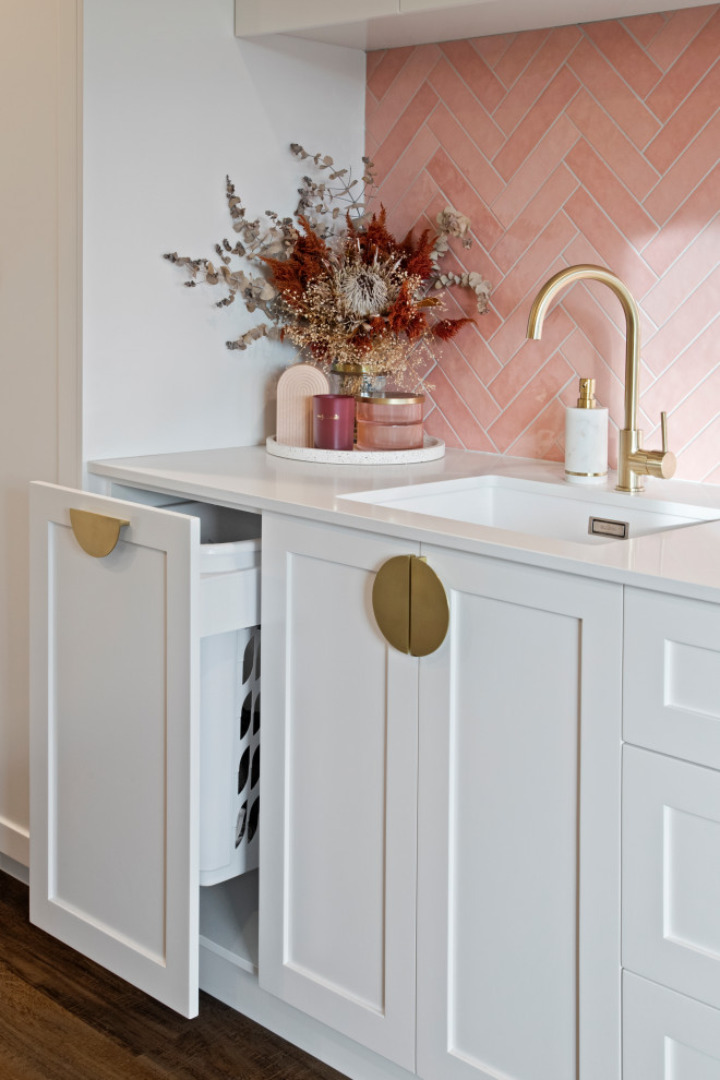 Mid-sized contemporary l-shaped utility room in Other with a single-bowl sink, shaker cabinets, white cabinets, pink splashback, mosaic tile splashback, white walls, light hardwood floors, a stacked washer and dryer, brown floor and white benchtop.