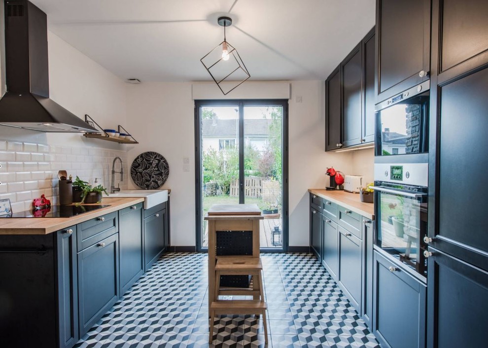 Design ideas for a mid-sized transitional galley kitchen in Nantes with a farmhouse sink, black cabinets, wood benchtops, subway tile splashback, cement tiles, with island, recessed-panel cabinets, white splashback, stainless steel appliances, multi-coloured floor and brown benchtop.