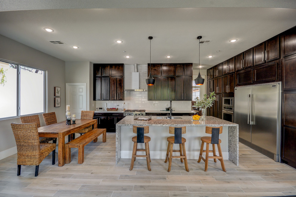 Mid-sized transitional l-shaped open plan kitchen in Las Vegas with a farmhouse sink, shaker cabinets, dark wood cabinets, granite benchtops, white splashback, porcelain splashback, stainless steel appliances, porcelain floors, with island and grey floor.
