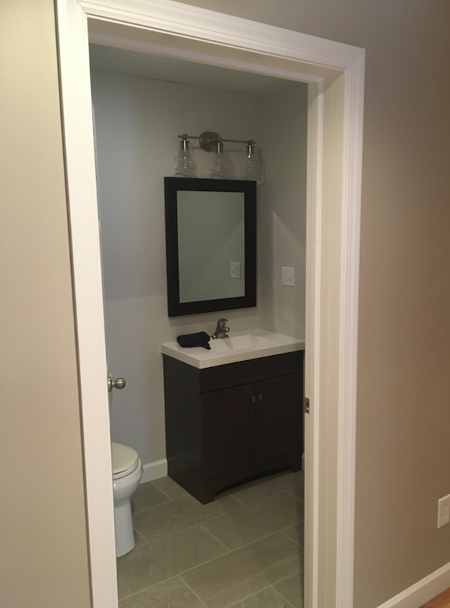 Small transitional laminate floor and beige floor powder room photo with flat-panel cabinets, medium tone wood cabinets, a one-piece toilet, gray walls, an integrated sink and solid surface countertops