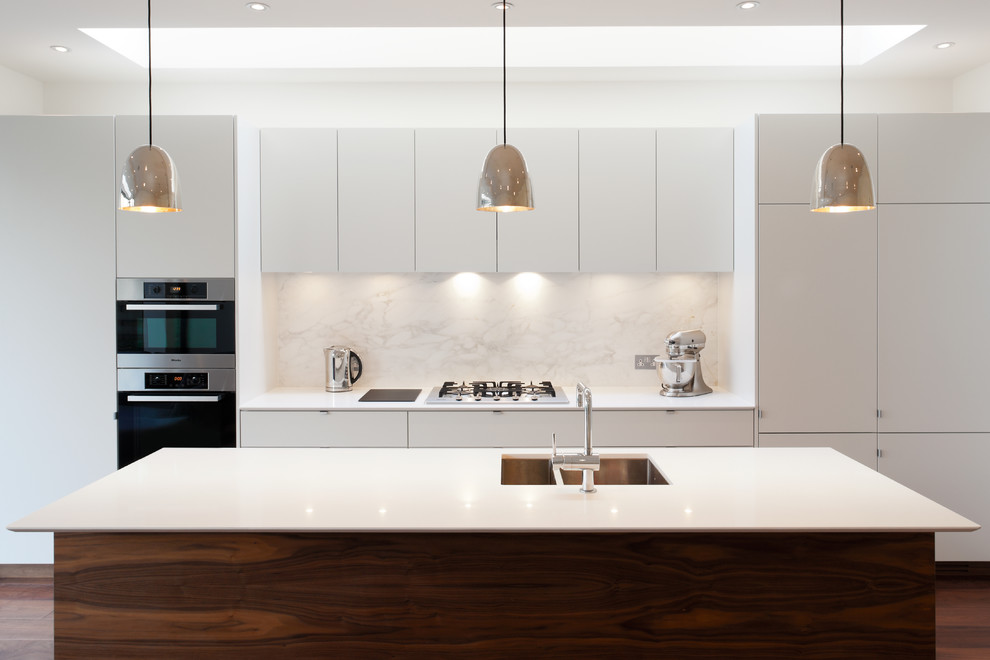 This is an example of a mid-sized contemporary single-wall eat-in kitchen in London with an integrated sink, flat-panel cabinets, white cabinets, solid surface benchtops, white splashback, stone slab splashback, panelled appliances, medium hardwood floors, with island and brown floor.