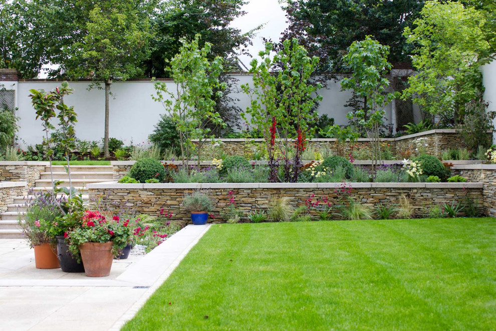 Modelo de jardín moderno de tamaño medio en verano en patio trasero con jardín francés, muro de contención, exposición total al sol, adoquines de piedra natural y con piedra