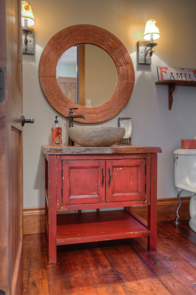 Mid-sized country powder room in Other with furniture-like cabinets, red cabinets, a two-piece toilet, grey walls, medium hardwood floors, a vessel sink, brown floor and brown benchtops.