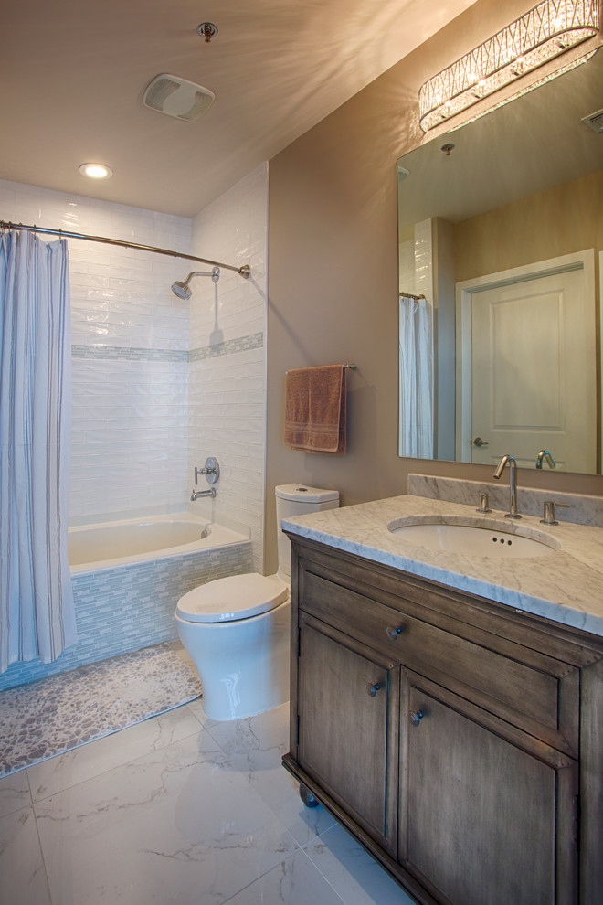 Photo of a mid-sized contemporary master bathroom in Nashville with a drop-in sink, furniture-like cabinets, medium wood cabinets, marble benchtops, an alcove tub, a shower/bathtub combo, a one-piece toilet, white tile, ceramic tile, beige walls and marble floors.