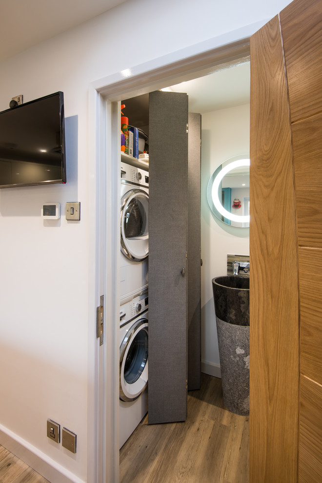This is an example of a small contemporary single-wall utility room in West Midlands with a single-bowl sink, beige walls, medium hardwood floors, a stacked washer and dryer and brown floor.
