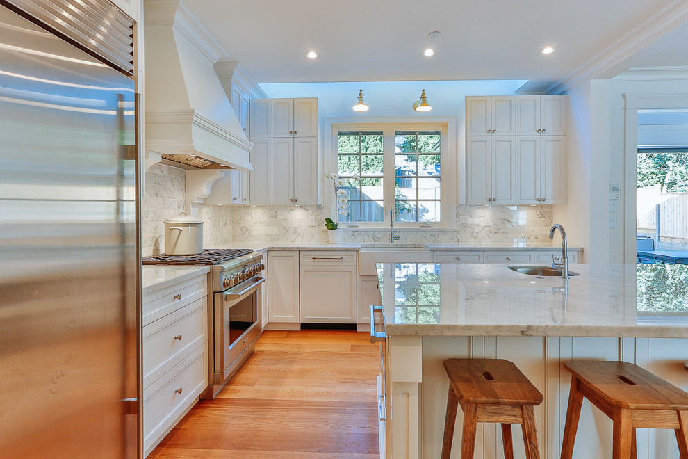 Photo of a mid-sized contemporary u-shaped eat-in kitchen in Vancouver with a farmhouse sink, shaker cabinets, white cabinets, marble benchtops, grey splashback, stone tile splashback, stainless steel appliances, medium hardwood floors and with island.