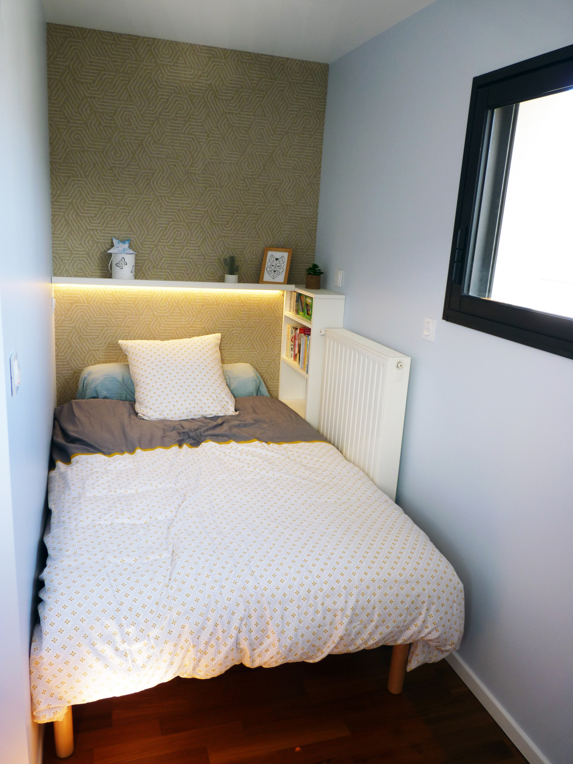 Petit coin lit avec un bandeau LED en chevet. L'espace a été optimisé pour qu'un lit 120cm passe avec la contrainte de ne pas bouger le radiateur. Une petite bibliothèque et une tablette en font un en