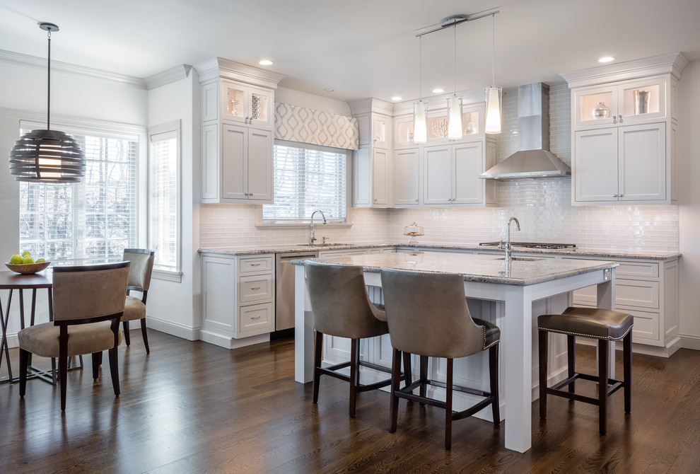 Expansive transitional l-shaped open plan kitchen in New York with a drop-in sink, recessed-panel cabinets, white cabinets, white splashback, stainless steel appliances, medium hardwood floors, with island, brown floor, grey benchtop, granite benchtops and subway tile splashback.