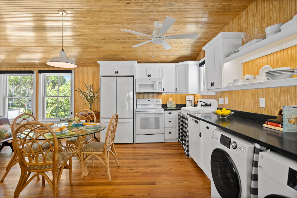 Design ideas for a small beach style l-shaped open plan kitchen in Tampa with dark hardwood floors, brown floor, wood, a farmhouse sink, beaded inset cabinets, white cabinets, brown splashback, timber splashback, white appliances, no island and black benchtop.