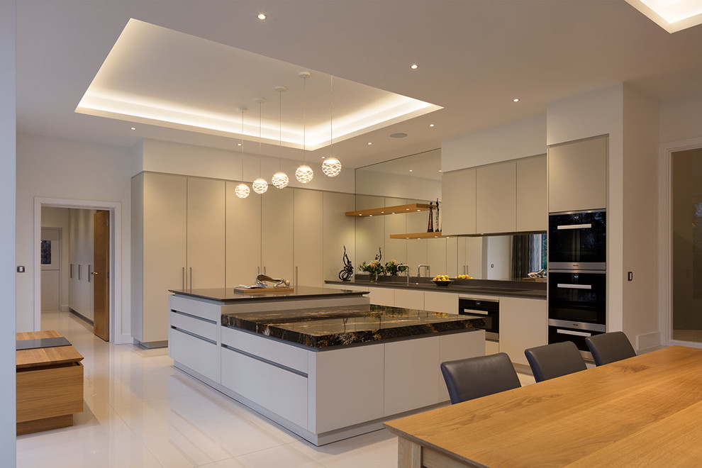 This is an example of an expansive contemporary l-shaped open plan kitchen in London with flat-panel cabinets, beige cabinets, granite benchtops, metallic splashback, mirror splashback, with island, white floor, black benchtop and black appliances.
