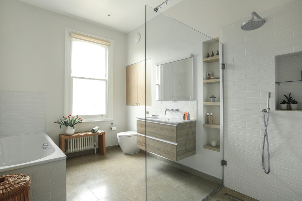 This is an example of a mid-sized contemporary master bathroom in London with flat-panel cabinets, medium wood cabinets, a drop-in tub, a curbless shower, a one-piece toilet, white tile, ceramic tile, white walls, limestone floors, green floor, an open shower, white benchtops and a console sink.