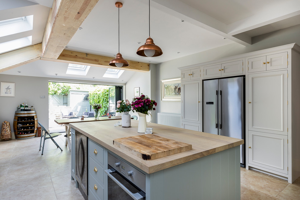 This is an example of a mid-sized country l-shaped eat-in kitchen in London with a farmhouse sink, shaker cabinets, white cabinets, wood benchtops, stainless steel appliances, limestone floors, with island and beige floor.