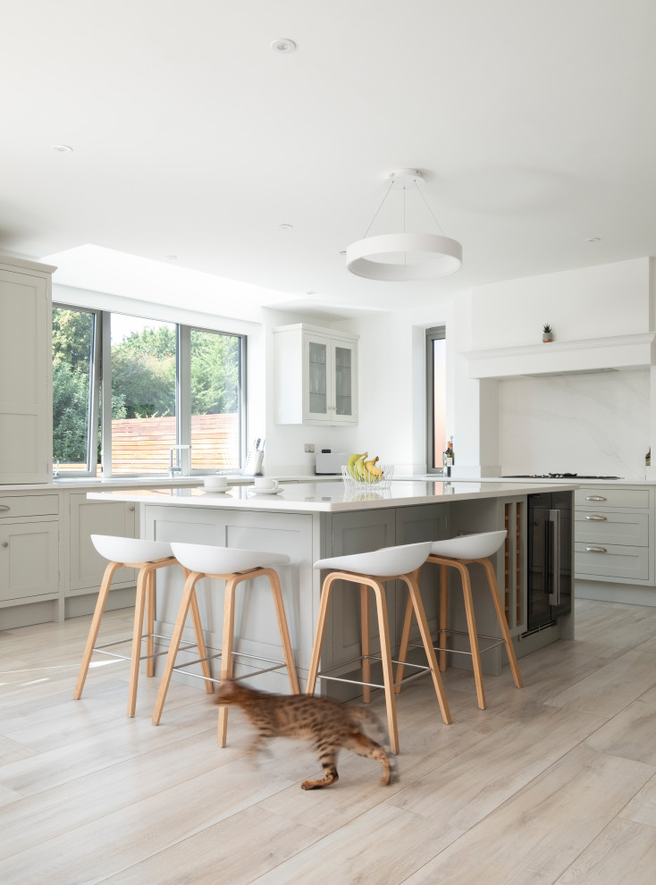 Design ideas for a large classic u-shaped open plan kitchen in Cardiff with a built-in sink, shaker cabinets, grey cabinets, quartz worktops, white splashback, engineered quartz splashback, stainless steel appliances, light hardwood flooring, an island, white worktops and a chimney breast.