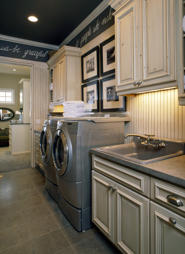 Design ideas for a mid-sized traditional single-wall dedicated laundry room in Denver with a drop-in sink, a side-by-side washer and dryer, grey floor, raised-panel cabinets, distressed cabinets, concrete benchtops, ceramic floors and black walls.