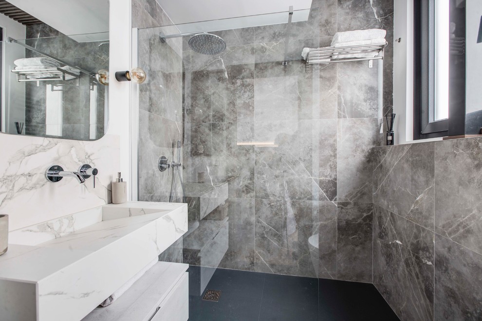 This is an example of a small industrial 3/4 bathroom in Madrid with a wall-mount toilet, gray tile, marble, grey walls, ceramic floors, a trough sink, engineered quartz benchtops, black floor, an open shower, flat-panel cabinets, white cabinets, an alcove shower and beige benchtops.